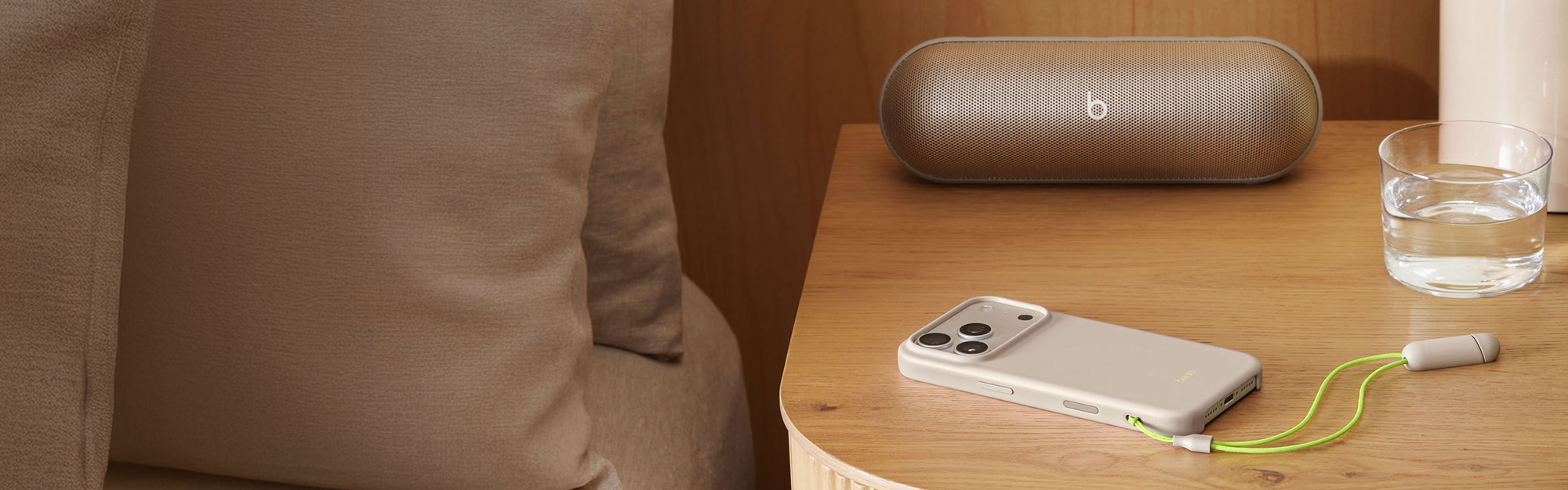 Beats Pill in Champagne Gold and  Beats iPhone 17 Pro Kickstand Case in Lime Stone on top of a night stand next to a cup of water.