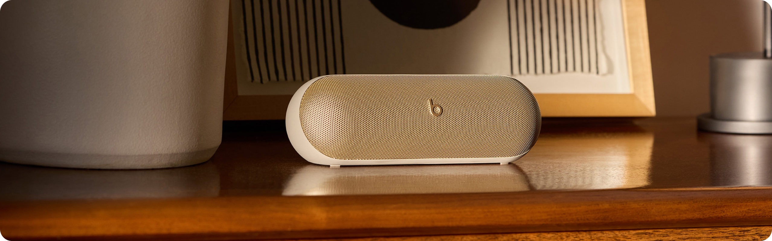 Gold Beats Pill speaker sits on a wooden dresser, with an abstract black and white framed artwork in the background.